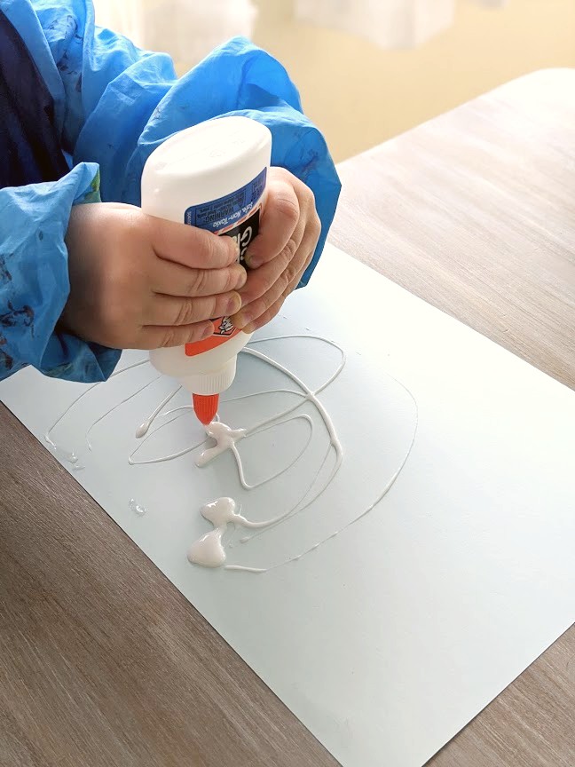 Young boy squeezing a tube of glue onto a piece of white cardstock to make a design for his salt art painting. 